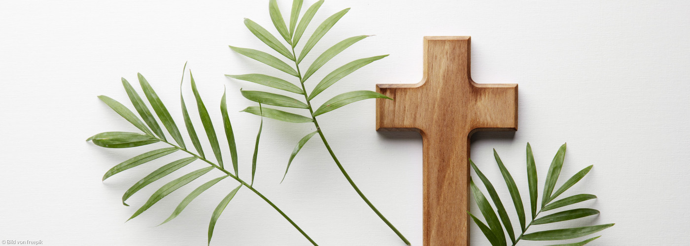 Ein Holzkreuz auf weißem Untergrund. Daneben liegen drei Palmblätter