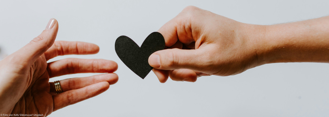 Zwei Häne. Eine Hand reicht ein Papierherz einer anedren geöffneten zum Empfang bereiten Hand 